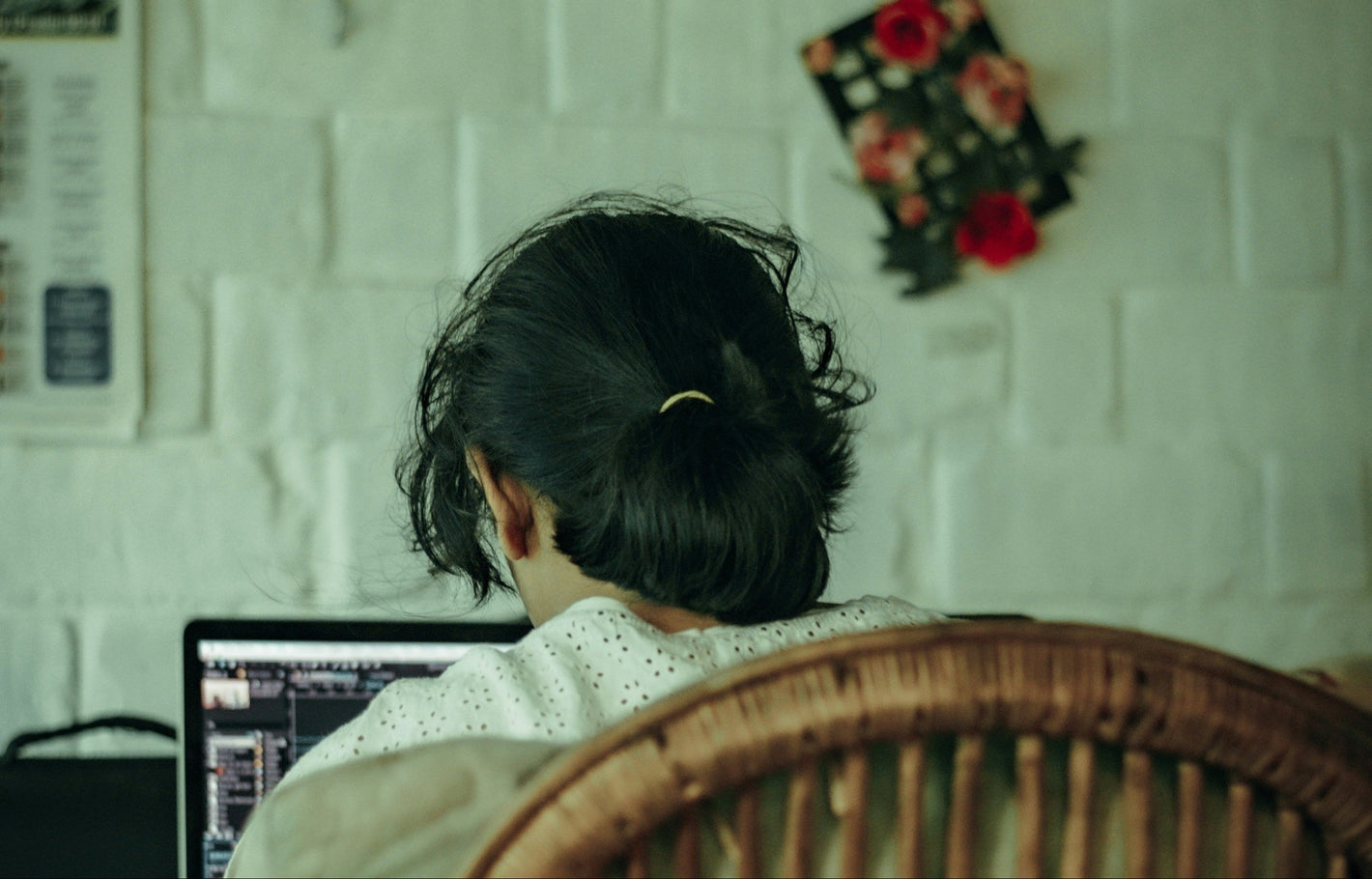 Person sitting on a wicker chair using a laptop against a white brick wall.