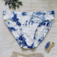 Blue and white tie-dye underwear on a light wooden surface with a plant and small bottle.