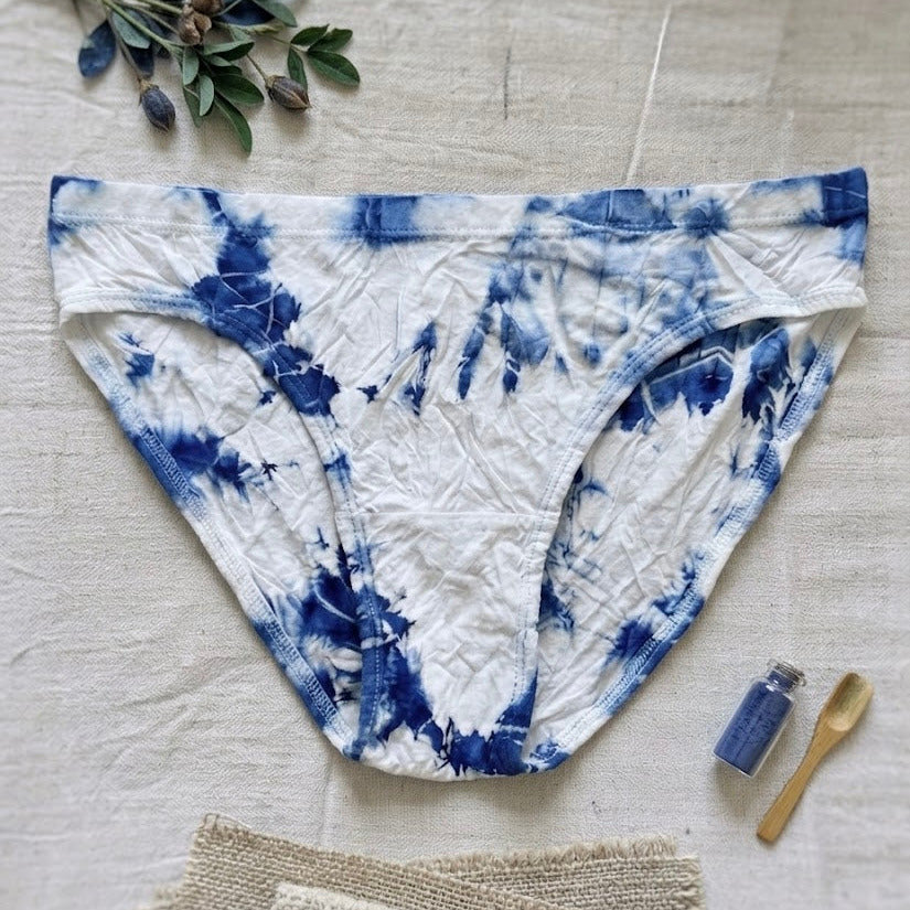 Blue and white tie-dye underwear on a light wooden surface with a plant and small bottle.