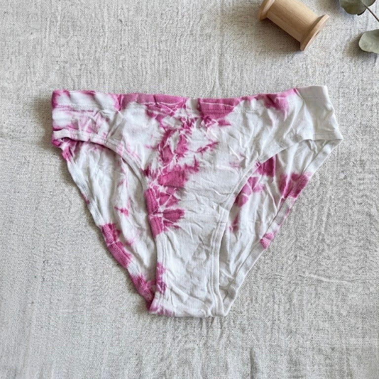 Pink and white tie-dye underwear on a textured surface with a wooden spool and green leaves.